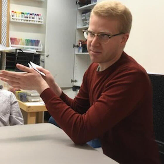 Ethan Cohen in a red shirt sitting at a desk speaking to someone off camera