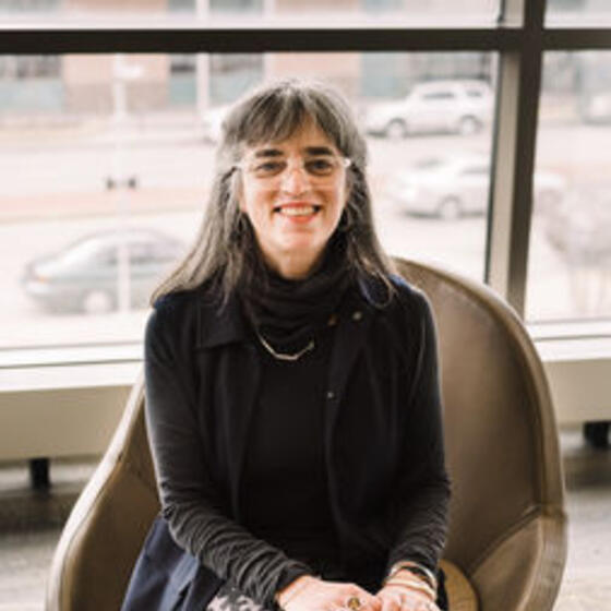 Laura Levitt sitting in a chair with a black shirt and colorful skirt on smiling and staring into the camera