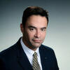 Hiram Aldarondo in a black suit with a a black and gold tie staring into the camera 