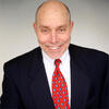 Christopher Swann wearing a black suit and red tie smiling and staring into the camera