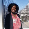 Meaonka Hadden standing at a window in a pink shirt and black suit coat smiling into the camera