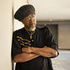 Kimani Nehusi wearing a black shirt and hat. His arms are crossed and he is staring off.