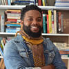 Michael Wilson in a jean jacket with a yellow scarf in front of a book shelf smiling into the camera