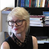 image of Olia sitting in front of a bookcase wearing black glasses and looking at the camera