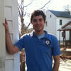 Thomas McKeon in a blue shirt standing outside with his hand on the side of a building smiling into the camera