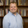 Ryan Sentner in a blue shirt standing in front of a book case smiling and looking into the camera