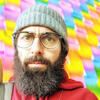 Jordan Shapiro in a winter hat and red shirt staring into a camera behind a multi colored wall