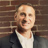 Kevin M. Glass in a black suit coat standing in front of a brick wall smiling into the camera